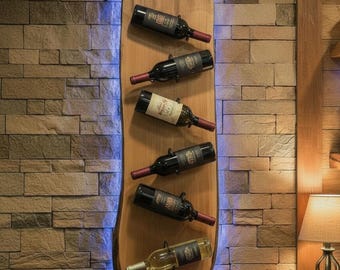 Puede incluir: Un botellero de madera con diseño de borde natural, que contiene seis botellas de vino y cuatro copas de vino. El botellero está montado en una pared de piedra e iluminado con luz azul. Las botellas son oscuras con corchos rojos.