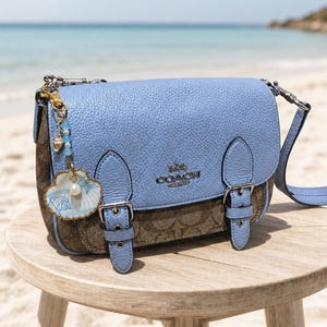 May include: A light blue and brown monogram print leather shoulder bag with a front flap, two buckle straps, and a silver-tone Coach logo. A decorative charm featuring a small seashell, blue beads, and a pearl hangs from the side hardware.