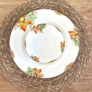 Könnte beinhalten: Weißes Keramik-Teller- und Schüssel-Set mit Goldrand und farbenfrohem Blattdesign. Die Blätter sind in Gelb-, Orange- und Grüntönen gehalten. Das Set steht auf einem runden, gewebten Platzset.