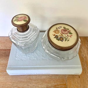 Könnte beinhalten: Eine klare, facettierte Glasparfümflasche und ein passendes Glas mit floralen Deckeln. Die Deckel haben einen cremefarbenen Hintergrund mit rosa und gelben Rosen. Die Artikel liegen auf einem hellblauen Buch mit dem Titel "Jane Eyre".