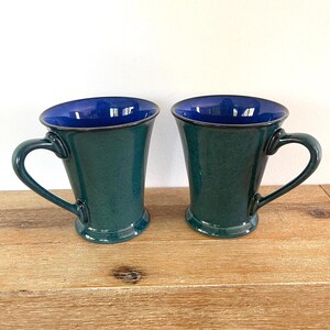 May include: Two ceramic mugs with a teal exterior and a deep blue interior. The mugs have a curved handle and a flared shape. The mugs are displayed on a wooden surface.
