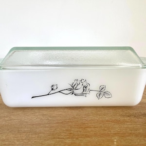 May include: White rectangular casserole dish with a clear glass lid. A black rose design is on the front of the dish. The dish is on a wooden surface.
