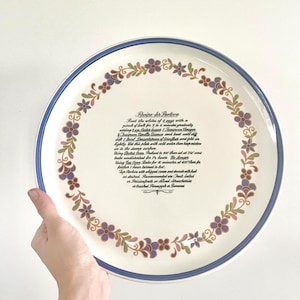 May include: A circular ceramic serving plate with a blue border and a floral pattern in purple, green, and brown. The plate features a recipe for Pavlova in black text.