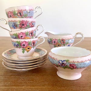 May include: A vintage tea set with pink teacups, saucers, a creamer, and a sugar bowl. Each item is decorated with a floral design in pink, blue, and yellow, with gold edging. The set includes six teacups, six saucers, a creamer, and a sugar bowl.