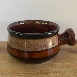 May include: A ceramic soup bowl with a handle. The bowl has a dark brown interior and a black rim. The exterior features bands of brown and tan colors. The bowl is sitting on a wooden surface.