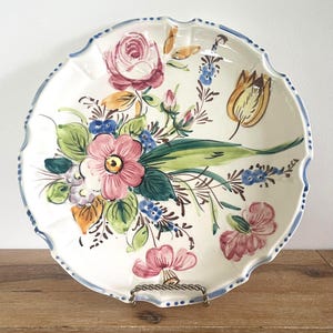 May include: A white ceramic serving plate with a scalloped edge and a hand-painted floral design. The flowers are pink, red, yellow, and blue, with green leaves. The rim has a blue border. The plate is on a metal stand.