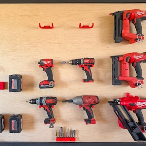 May include: A light-colored wooden wall-mounted organizer board featuring red plastic mounts holding various red and black Milwaukee power tools, including three nail guns, two drills, an impact driver, four battery packs, and several drill bits.