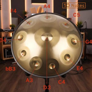 May include: A golden steel tongue drum with 10 notes labeled. The instrument has a central indentation and several circular depressions around the surface. The drum is mounted on a black stand, with a wooden background.