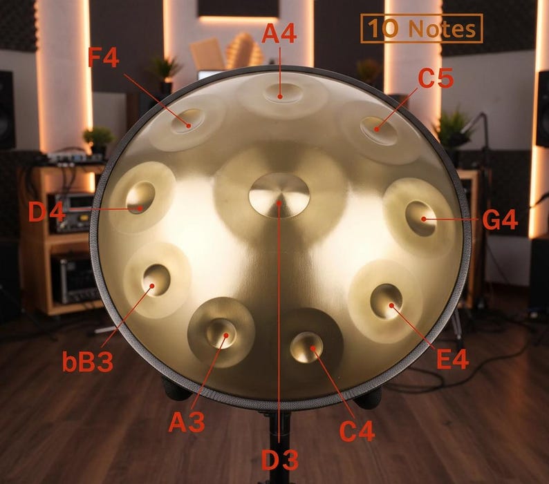 May include: A golden steel tongue drum with 10 notes labeled. The instrument has a central indentation and several circular depressions around the surface. The drum is mounted on a black stand, with a wooden background.