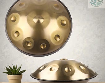 以下が含まれることがあります： 中央のドームと周囲に配置された8つのトーンフィールドを備えた金色のスチール製ハンドパン。楽器は滑らかな金属仕上げと暗い縁を持ち、上からの視点と側面からの視点の両方で表示されています。