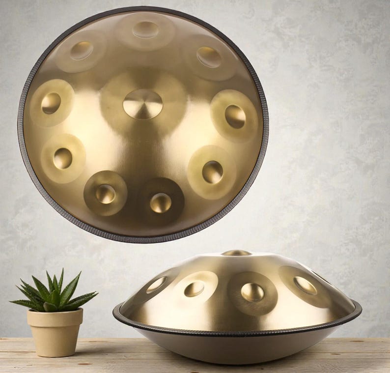 May include: A golden-colored steel tongue drum with a dark rim. The instrument has multiple circular indentations on its surface. A small potted succulent plant sits on a wooden surface in the foreground.