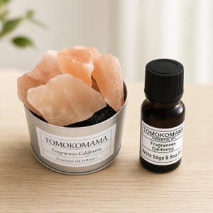 以下が含まれることがあります： ピンク色のヒマラヤ岩塩が入った金属製の容器で、「TOMOKOMAMA Fragrances California Essential Oil Diffuser」と表示されています。その隣には、黒いキャップが付いた小さなエッセンシャルオイルのボトルがあり、「TOMOKOMAMA Fragrances California White Sage & Sea Salt」と表示されています。