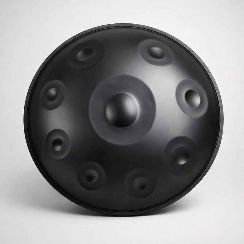 May include: A black steel handpan instrument with a rounded shape and multiple circular indentations. The instrument has a central dome and a smooth, matte finish. The handpan is designed for melodic percussion and is often used in meditative music.