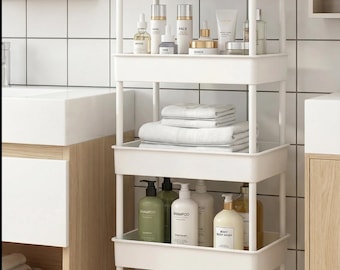 Puede incluir: Un carrito blanco con ruedas y cuatro estantes, lleno de artículos de baño. El estante superior contiene una pequeña planta, frascos y objetos decorativos. El segundo estante tiene productos para el cuidado de la piel. El tercer estante tiene toallas blancas dobladas.
