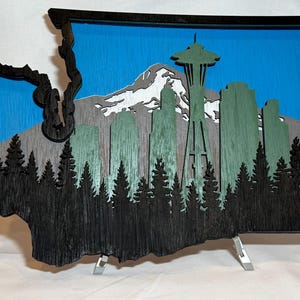 May include: A wooden wall art piece in the shape of Washington State, featuring a layered design. The artwork depicts a forest, mountains, the Seattle skyline, and the Space Needle against a blue sky. The frame is black.