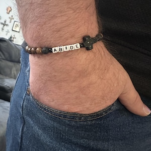 May include: A beaded bracelet with the word "ABIDE" spelled out in white letter beads. The bracelet features brown and black beads, and a cross-shaped charm. The bracelet is worn on a wrist.