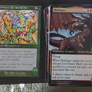 Könnte beinhalten: Zwei Magic: The Gathering Sammelkarten. Eine Karte zeigt eine farbenfrohe Illustration einer Kreatur namens "Sakura-Tribe Elder". Die andere Karte zeigt einen furchterregenden Dämonendrachen namens "Malfegor".