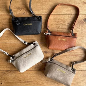 Puede incluir: Cuatro bolsos bandolera rectangulares pequeños con textura granulada, cada uno con cremallera superior, correa desmontable y el texto dorado GLYMMR impreso en la parte delantera. Los bolsos están disponibles en colores negro, tostado, blanco roto y gris claro.