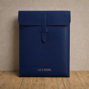 May include: A navy blue leather tablet sleeve with a flap and strap closure. The sleeve features a gold embossed logo reading "GLYMMR". The background is a neutral beige, and the sleeve rests on a wooden surface.