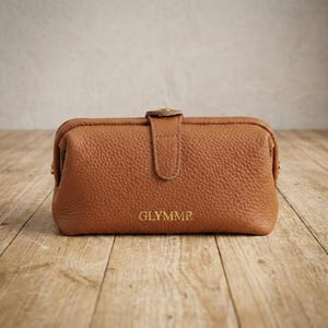 Puede incluir: Un estuche de cuero marrón con cierre de broche y la palabra "GLYMMR" grabada en la parte delantera. El estuche tiene una superficie texturizada y está sobre un fondo de madera.