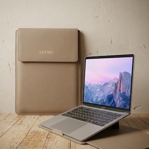 May include: A tan leather laptop sleeve with a flap closure and the gold-colored text "GLYMME". A silver laptop is open on a stand next to the sleeve. The laptop screen displays a mountain landscape.