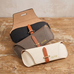 May include: Three leather eyeglass cases in black, gray, and cream. Each case has a contrasting brown strap with a gold-colored buckle. The word "GLYM" is printed in gold on the gray and cream cases. The cases are on a wooden surface.