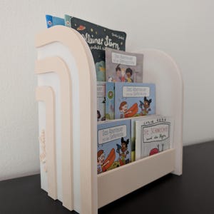 Könnte beinhalten: Ein weißes und hellpfirsichfarbenes Bücherregal aus Holz mit einem gestuften Bogendesign an den Seitenteilen. Das Regal enthält mehrere Kinderbücher mit Titeln wie Kleiner Stern, Das Abenteuer mit der Seifenkiste und Die Schnecke und der Regen.