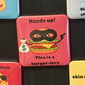 Peut inclure: Un aimant carré rose avec un hamburger de dessin animé portant un masque noir, tenant une arme et un sac d'argent. Le texte dit "Hands up!" et "This is a burger-lery."