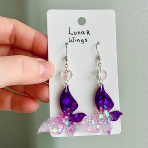 May include: A pair of handmade earrings featuring a butterfly design. The earrings have a purple and glittery wing design, with a clear bead and silver hook. The text "Luna R Wings" is printed on the white card.