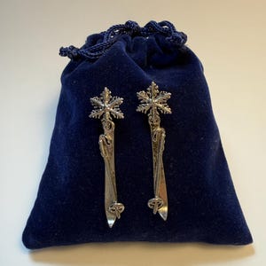 Puede incluir: Un par de pendientes de copo de nieve plateados con un diseño largo y delgado. Los pendientes se muestran frente a una bolsa de terciopelo azul oscuro con cierre de cordón. El diseño de copo de nieve está en la parte superior.