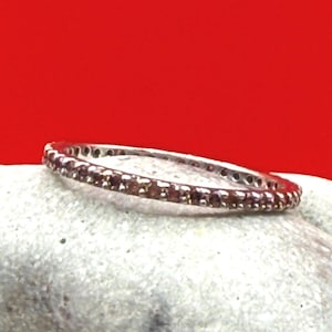 May include: A silver ring with a band of small, round, reddish-brown gemstones. The ring is set against a light gray surface with a red background. The gemstones are evenly spaced around the band.