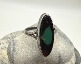 May include: Silver ring with an oval black stone and a green accent. The ring is set on a gray stone, showcasing the jewelry's design and color contrast. The ring's band is thin and delicate.