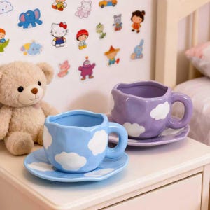 May include: Two ceramic teacups and saucers, one light blue and one lavender, each with white cloud designs. The cups have rounded handles and sit on matching saucers. A plush teddy bear and stickers are in the background.