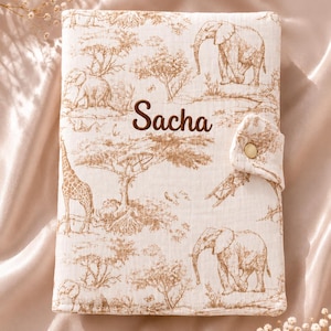 Peut inclure: Une couverture en tissu rectangulaire avec un fond beige clair et un imprimé de style toile de Jouy marron représentant des éléphants, des girafes et des arbres. Le prénom Sacha est brodé en marron sur le devant, doté d'un rabat avec bouton pression.