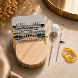 May include: A small metal darning loom tool with a round wooden base, featuring a series of silver hooks and yellow elastic bands. The set includes a long metal needle with a white spherical top and a separate metal hook tool for textile repair.
