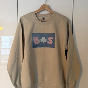 May include: A beige crew neck sweatshirt with long sleeves. The front displays a rectangular patch with the letters "B" and "S" in pink, and a green shamrock. The patch is set against a blue denim-like background. The sweatshirt is suspended on a wooden hanger.