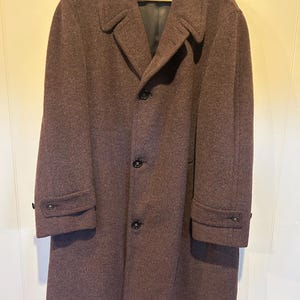 Puede incluir: Un abrigo largo de lana burdeos oscuro con un diseño clásico. El abrigo tiene un cuello con solapas, tres botones negros en la parte delantera y botones en los puños. Cuelga de una percha de madera.