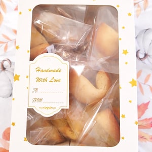 May include: A rectangular white box with a clear window filled with individually wrapped fortune cookies. The box features a yellow star pattern and a white label with gold trim that reads Handmade With Love, TO: ______, FROM: ______.