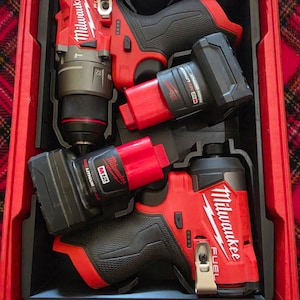May include: A red and black Milwaukee M12 Fuel power tool set inside a red carrying case. The set includes a drill driver and an impact driver, each with a black and red M12 Redlithium battery pack attached, featuring white Milwaukee branding.