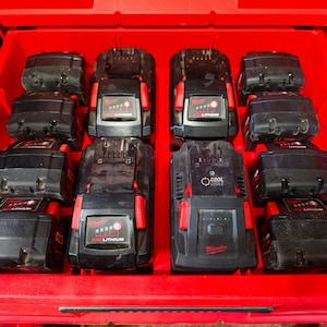 May include: A bright red plastic storage case containing ten black power tool batteries and one battery charger. The batteries feature red accents and the Milwaukee RedLithium logo. The charger is labeled with Cool Cycle text and Milwaukee branding.