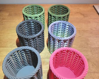 May include: Six small, woven baskets in various colors: two green, two gray, one pink, and one with blue and purple hues. The baskets are cylindrical with a textured, braided design. They sit on a wooden surface.