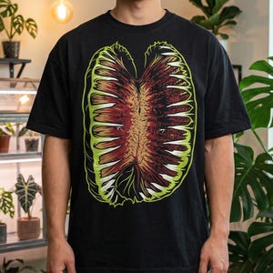 May include: Black t-shirt featuring a detailed, colorful graphic of a plant-like form. The design has a central brown area with red and white accents, outlined in green. The shirt is a classic crew neck style.