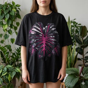 May include: Black t-shirt featuring a large, detailed graphic of a tropical leaf. The leaf design showcases a striking contrast of dark gray and vibrant pink hues, creating a bold visual statement. The shirt is a casual, oversized fit.