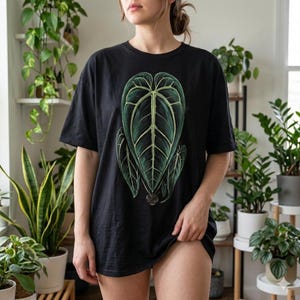 May include: Black t-shirt featuring a detailed illustration of a large, dark green leaf with lighter green veins. The leaf design is centered on the front of the shirt. The shirt is a short-sleeved, casual style.