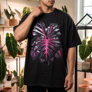 May include: Black t-shirt featuring a large, stylized leaf design with a vibrant pink center and dark gray outer details. The graphic is centered on the front of the shirt. The shirt is a relaxed fit.
