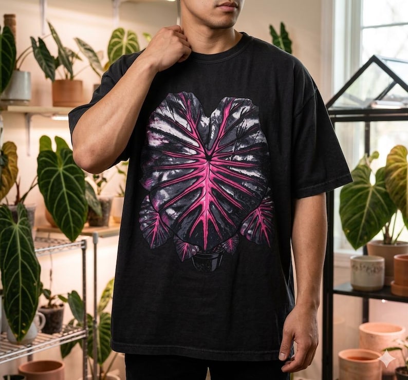 May include: Black t-shirt featuring a large, stylized leaf design with a vibrant pink center and dark gray outer details. The graphic is centered on the front of the shirt. The shirt is a relaxed fit.