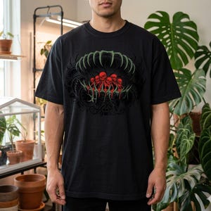 May include: Black t-shirt featuring a graphic design of a mouth-like shape with green teeth and red skulls. The design is centered on the front of the shirt. The shirt is made of a soft, dark fabric.