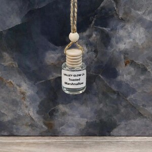 May include: A small, clear glass bottle with a wooden cap and a label that reads "VALLEY GLOW CA Toasted Marshmallow." The bottle is suspended by a beige string with a wooden bead. The background is a dark, marbled surface.
