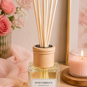 May include: A clear glass diffuser bottle with a wooden cap and reeds. The label reads "ROSÉ EMBRACE" with the @ValleyGlowCA handle. The image includes a lit candle, pink roses, and a pink scarf, creating a relaxing ambiance.