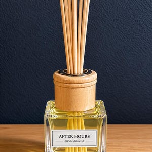 May include: A clear glass diffuser bottle with a wooden cap and reeds. The bottle contains a yellow liquid and a label that reads "AFTER HOURS @ValleyGlowCA". The background is a dark blue.